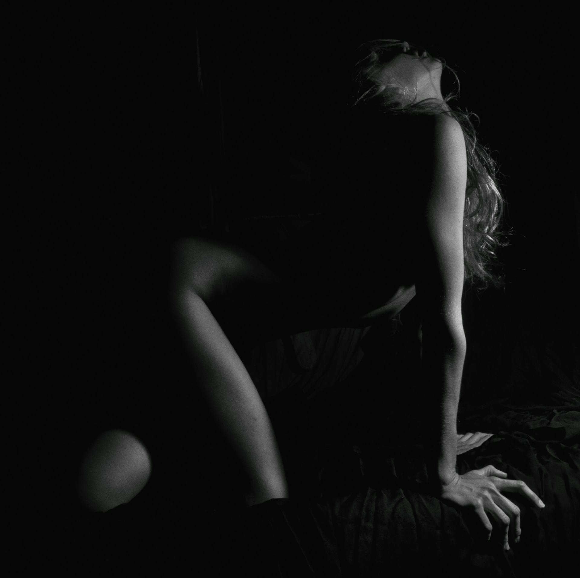 Photo en noir et blanc d'une femme à genoux sur un matelas au drap croisé, elle relève la tête avec satisfaction.