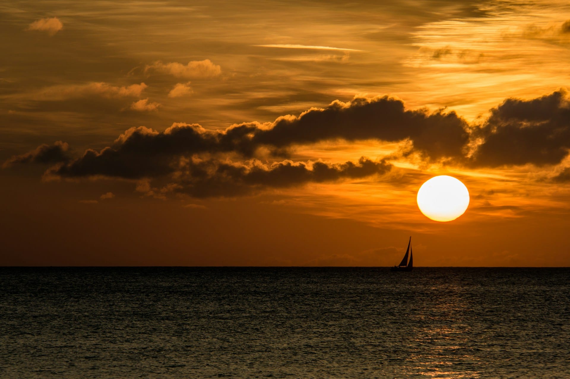 Sous un soleil couchant embrasant le ciel d'un orange vif, un voilier vient flirter avec le soleil rasant la mer.