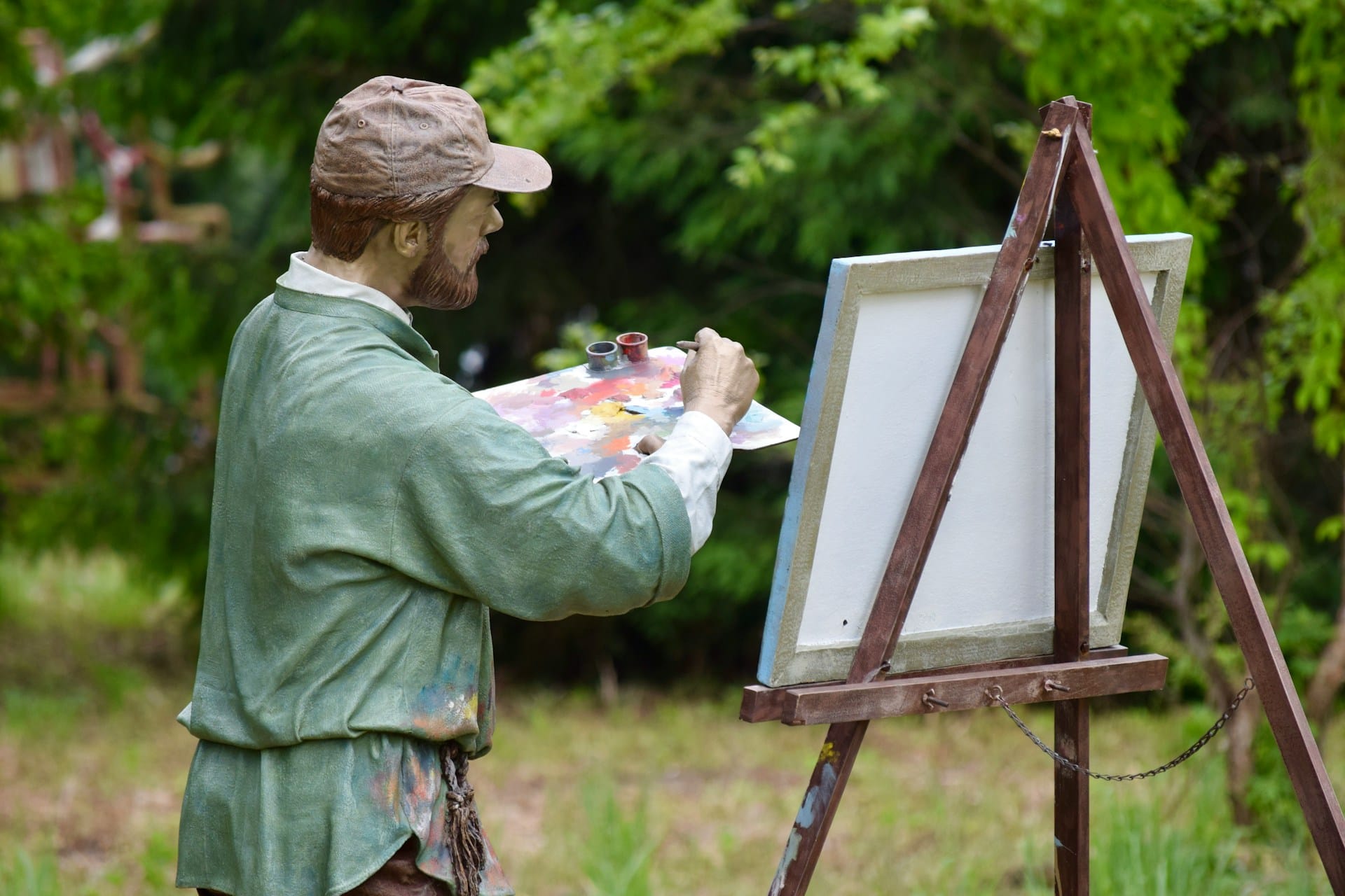 Une statue d'un peintre barbu devant son chevalet dans un jardin. Il prend de la peinture sur sa palette.