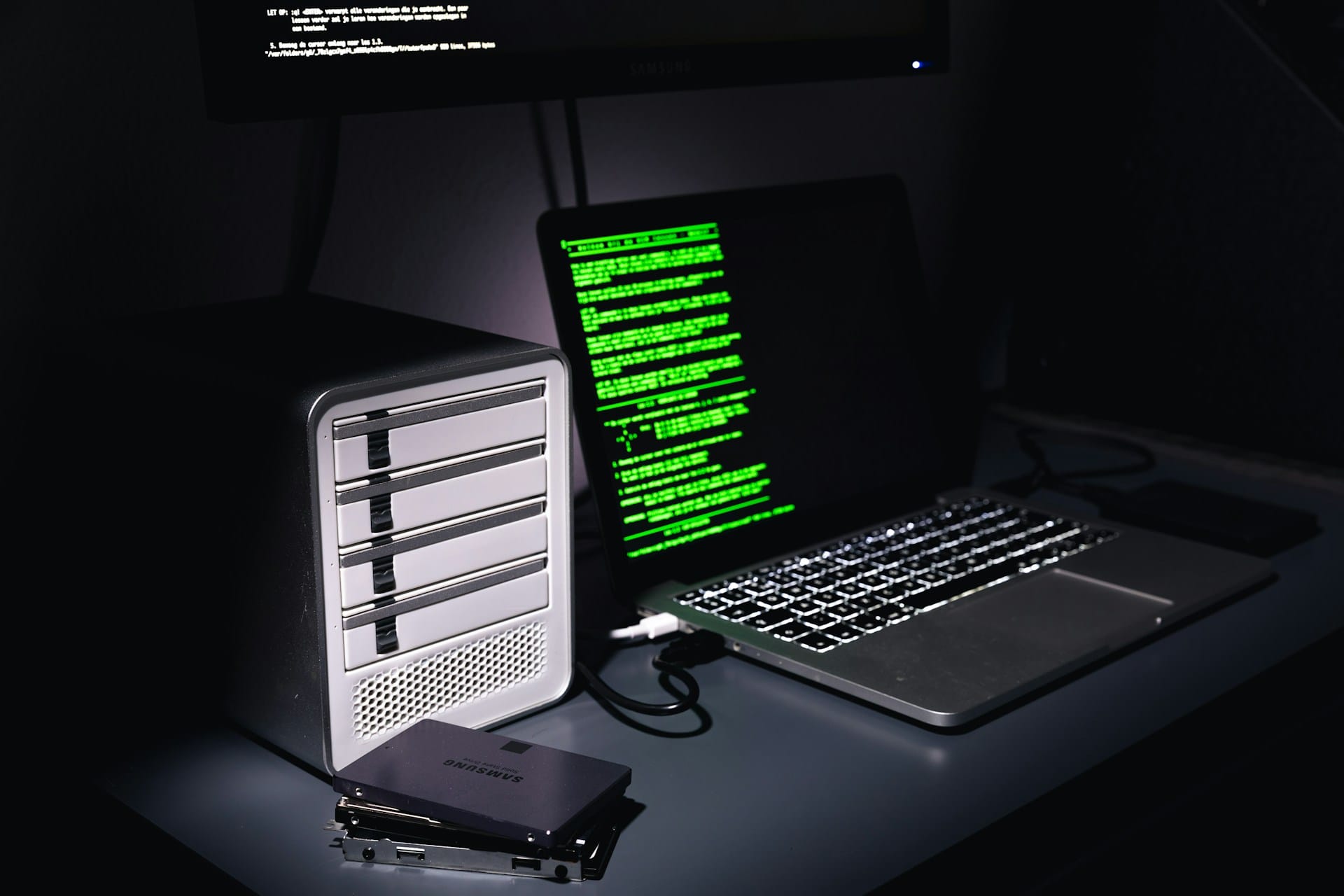 Sur un bureau dans le noir, l'écran d'un ordinateur portable laisse deviner des lignes d'un message écrit en vert. À côté du portable, des disques durs.