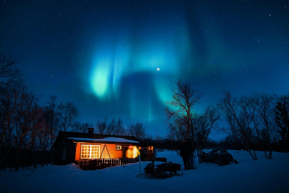 Une cabane en bois éclairée dans la nuit dans un paysage de neige. Au-dessus, une aurore boréale.
