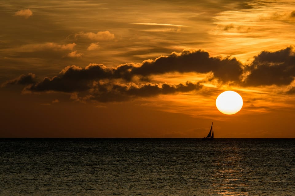 Sous un soleil couchant embrasant le ciel d'un orange vif, un voilier vient flirter avec le soleil rasant la mer.