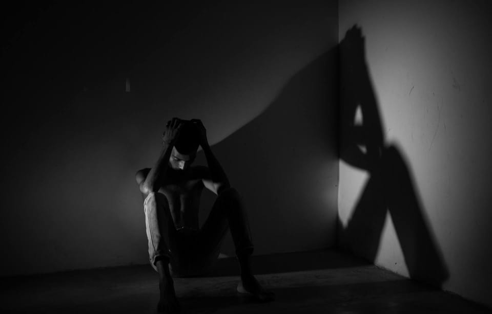 Photo en noir et blanc. Un homme recroquevillé dans un coin d'une pièce vide. Il se tient la tête entre les mains.