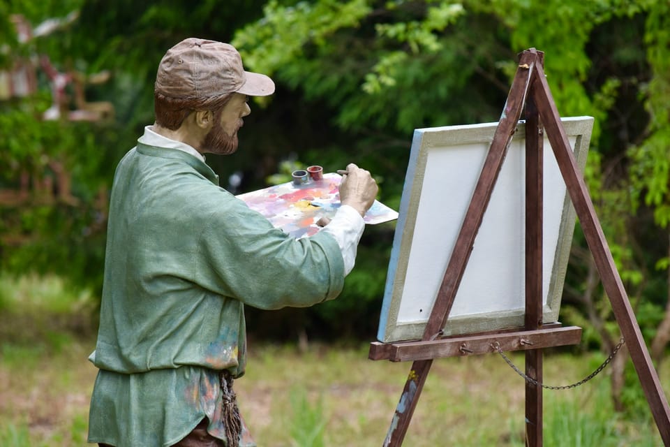 Une statue d'un peintre barbu devant son chevalet dans un jardin. Il prend de la peinture sur sa palette.