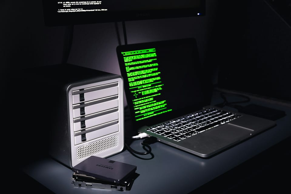 Sur un bureau dans le noir, l'écran d'un ordinateur portable laisse deviner des lignes d'un message écrit en vert. À côté du portable, des disques durs.