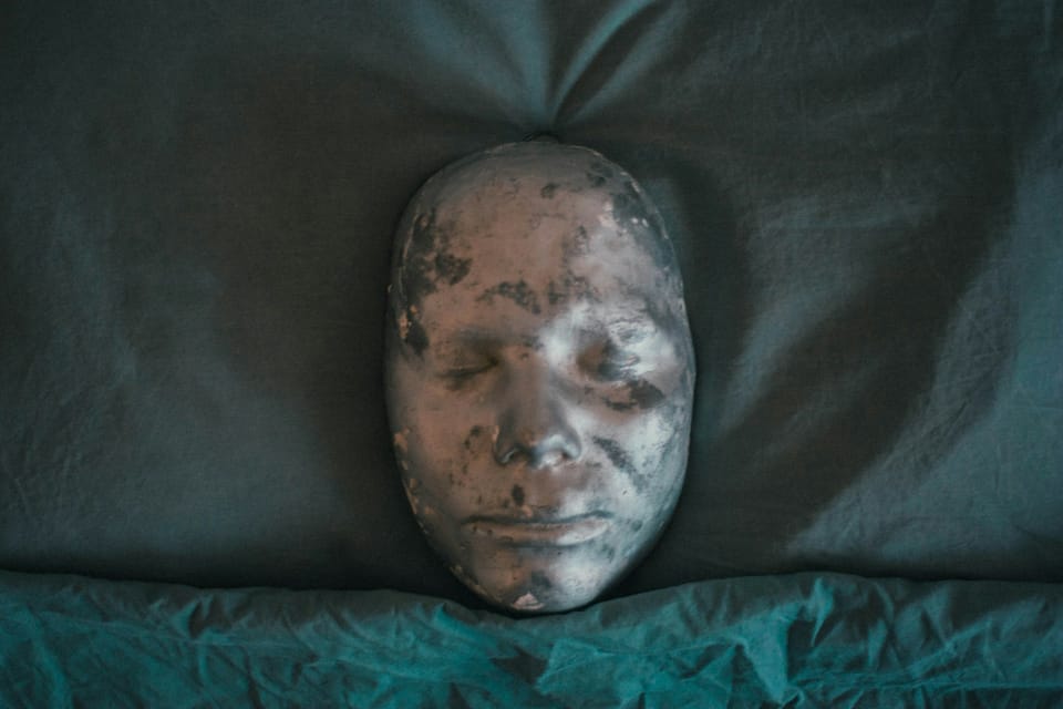 Un masque corrodé est posé sur un oreiller, à la frontière avec le drap.