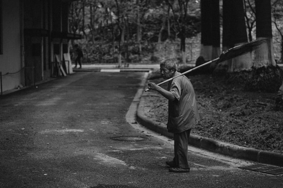 Sur une petite rue, un vieil homme au dos courbé porte un balais de paille.